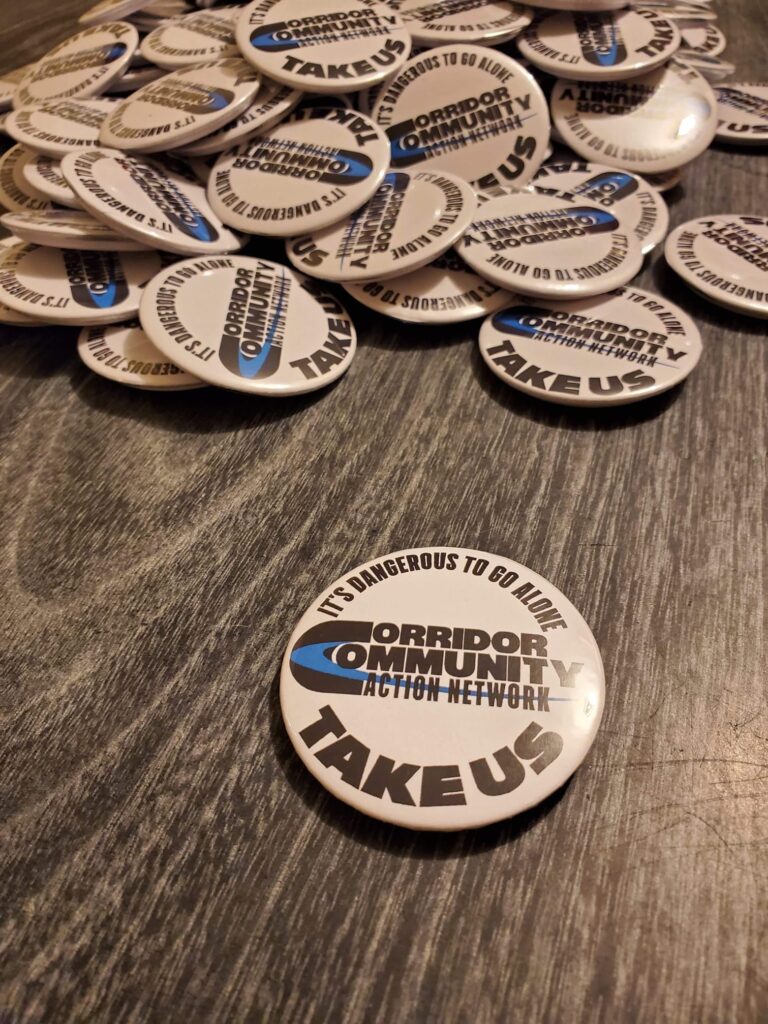 Button on a table that reads "It's dangerous to go alone, take us. Corridor Community Action Network."